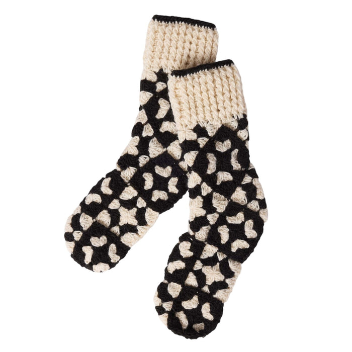 Wearing cozy MERSEA - LA PAZ CROCHET SLIPPER SOCKS, a person sits on the floor with a matching black and white crocheted hood, scarf, and gloves, paired with blue jeans and a black long-sleeve shirt, smiling softly at the camera.