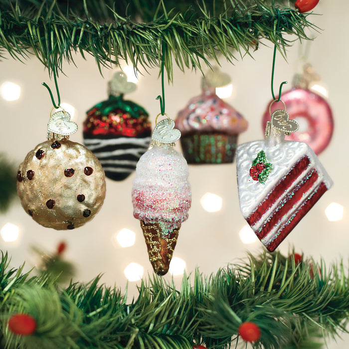 Close-up of a Christmas tree decorated with dessert-themed ornaments, including a cookie, ice cream cone, cupcakes, and a slice of cake, with festive lights blurred in the background.