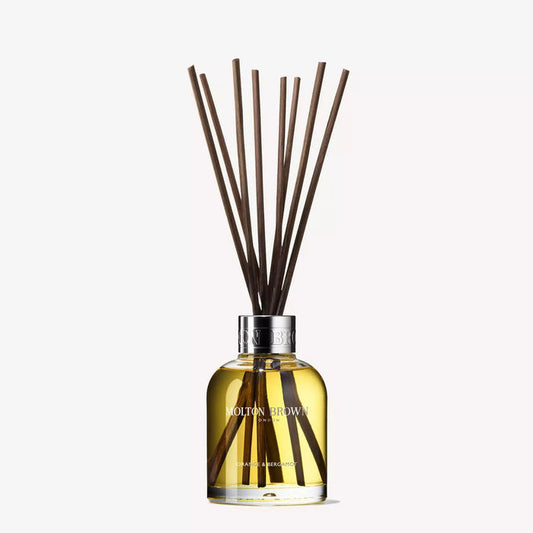 A clear glass bottle labeled "MOLTON BROWN - ORANGE AND BERGAMOT DIFFUSER" contains yellow liquid and several reed sticks, gently dispersing a citrus scent, set against a plain white background.