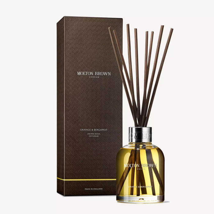 A clear glass bottle labeled "MOLTON BROWN - ORANGE AND BERGAMOT DIFFUSER" contains yellow liquid and several reed sticks, gently dispersing a citrus scent, set against a plain white background.