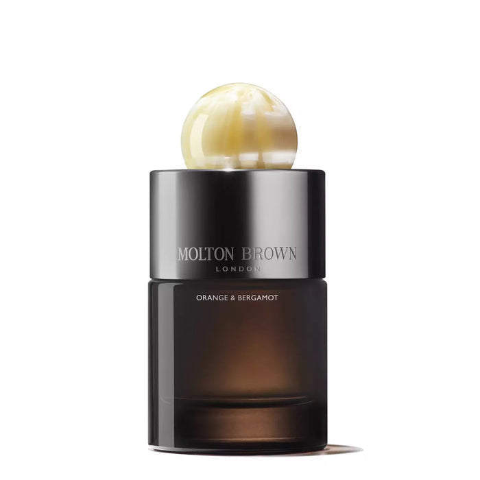 A bottle of MOLTON BROWN - ORANGE AND BERGAMOT EAU DE PARFUM with a dark cylindrical base and round marbled pearl-like cap, placed on a white background accented by orange flowers.