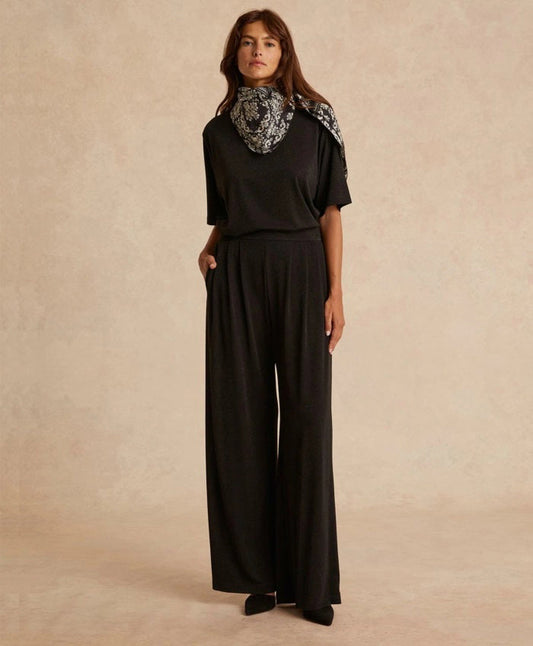 A woman stands against a neutral background wearing MOMONI Lanzarote Pant in Lurex Jersey, a loose black top, black shoes, and a black-and-white patterned scarf, with her hands in her pockets.