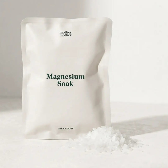 Two hands are cupped together holding a pile of white, flat, flaky pieces that resemble large grains of sea salt or wax flakes—these are MOTHER MOTHER - MAGNESIUM BATH SOAK 3OZ pure magnesium chloride flakes from MOTHER MOTHER, ideal for a restorative bath. The background is out of focus and neutral in color.