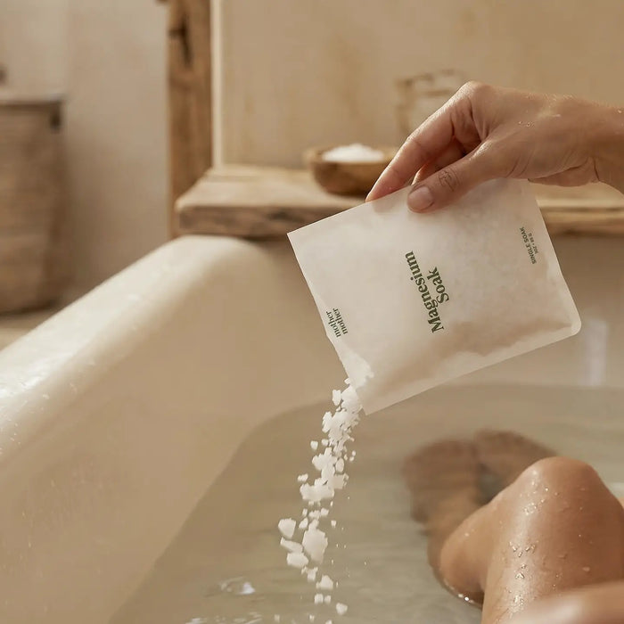 A white packet labeled “MOTHER MOTHER - MAGNESIUM BATH SOAK 3OZ” from the brand MOTHER MOTHER stands upright on a light surface, with a small pile of pure white magnesium chloride flakes—sourced from Zechstein magnesium—in front of it. The minimalist packaging features green text and simple design, making it perfect for a restorative bath experience.