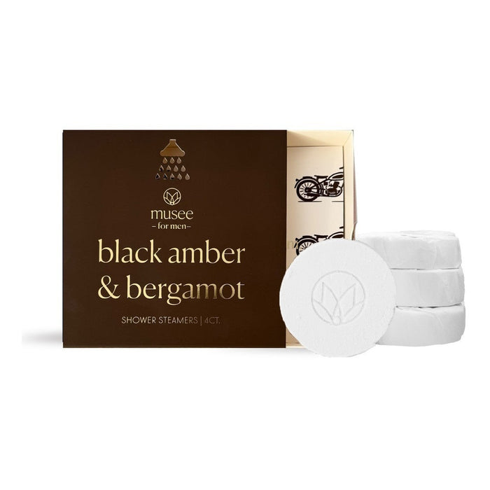 A brown box labeled “MUSEE - BLACK AMBER & BERGAMOT SHOWER STEAMERS” from the brand MUSEE is shown, with three white round steamers stacked in front. The partially open box reveals its interior and a motorcycle graphic, highlighting these natural shower aromatherapy products.