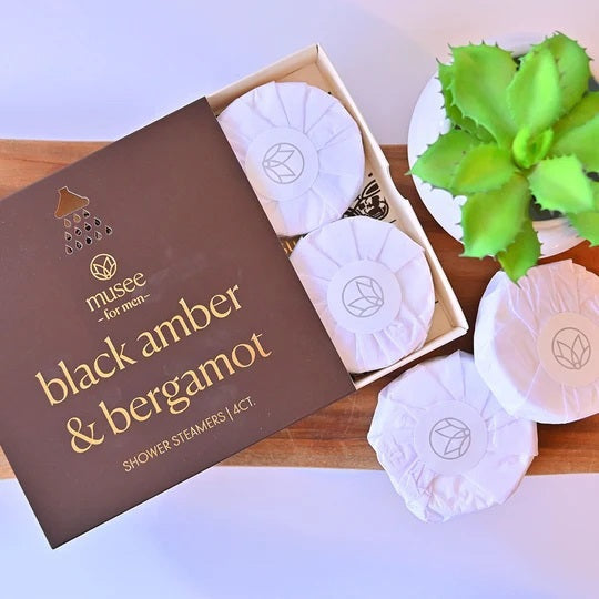 A brown box labeled “MUSEE - BLACK AMBER & BERGAMOT SHOWER STEAMERS” from the brand MUSEE is shown, with three white round steamers stacked in front. The partially open box reveals its interior and a motorcycle graphic, highlighting these natural shower aromatherapy products.