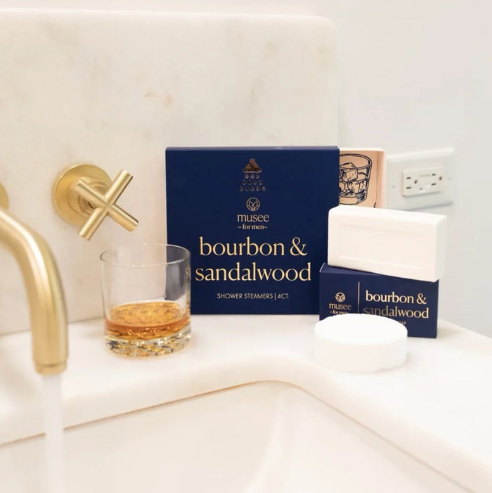 A blue box labeled “MUSEE - BOURBON & SANDALWOOD SHOWER STEAMERS” by MUSEE sits beside four white round steamers and an open lid adorned with a drawing of a whiskey glass, offering an indulgent aromatherapy shower experience enhanced with sandalwood essential oil.