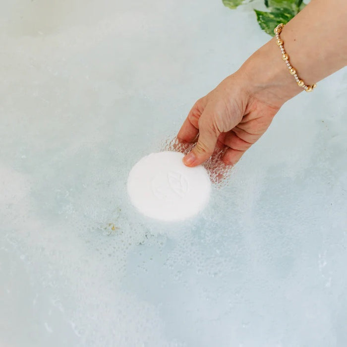A light blue cylindrical container labeled “MUSEE - CALM BATH TABLETS” by MUSEE is positioned next to a stack of six white, round bath tablets. The container highlights the “Water Lily & Linen” scent, evoking the tranquil atmosphere of natural bath products ideal for a soothing soak.