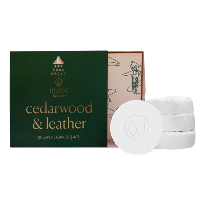 An open green box labeled “MUSEE - CEDARWOOD & LEATHER SHOWER STEAMERS” by MUSEE reveals four round white steamers stacked nearby and inner packaging with illustrations. Each steamer is infused with cedarwood essential oil, transforming your shower into a spa-like retreat.