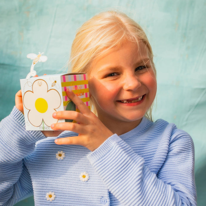 A light blue MUSEE - DAISY BATH BALM by MUSEE rests beside a beautifully decorated box featuring a white daisy with a yellow center and playful cartoon bees, including one bee attached on a wire. This delightful set is perfect for anyone who loves natural bath products and enjoys indulging in soothing bath balms.