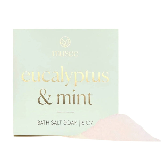 A square package labeled “MUSEE - EUCALYPTUS & MINT MINI BATH SALT SOAK” rests on a tray beside a green succulent, inviting you to enjoy the refreshing aroma of eucalyptus and mint with natural bath products from MUSEE.