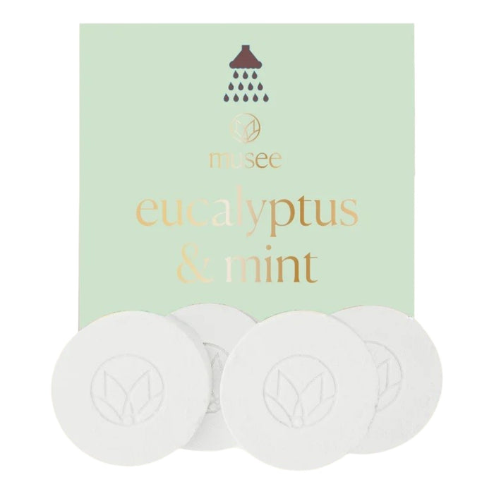 A pastel green packet labeled “MUSEE - EUCALYPTUS & MINT SHOWER STEAMERS” from MUSEE, featuring a shower icon, is displayed behind four round white shower steamers embossed with a leaf design—offering a spa-like experience with these natural bath products.