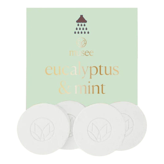 A pastel green packet labeled “MUSEE - EUCALYPTUS & MINT SHOWER STEAMERS” from MUSEE, featuring a shower icon, is displayed behind four round white shower steamers embossed with a leaf design—offering a spa-like experience with these natural bath products.