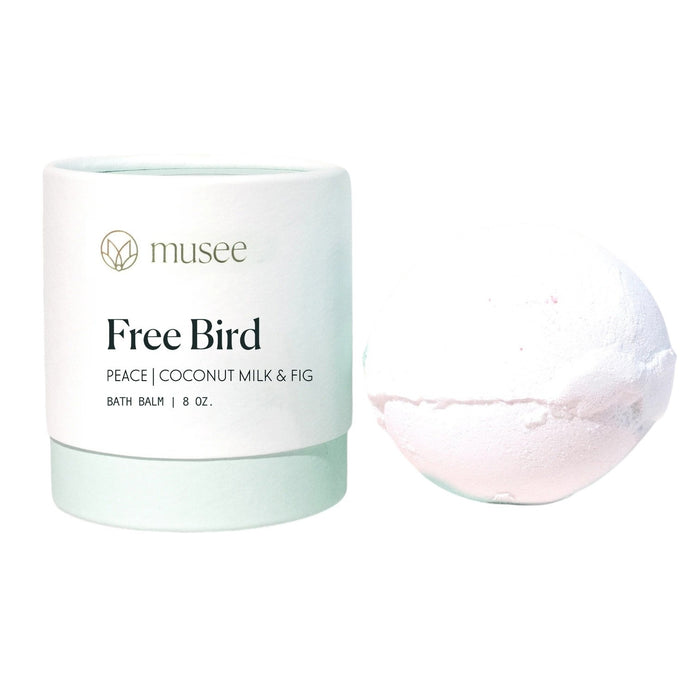 A light green and white container labeled “MUSEE - FREE BIRD THERAPY BATH BALM” sits beside a white bath bomb, offering a soothing and therapeutic bath experience with natural ingredients and coconut essential oil.