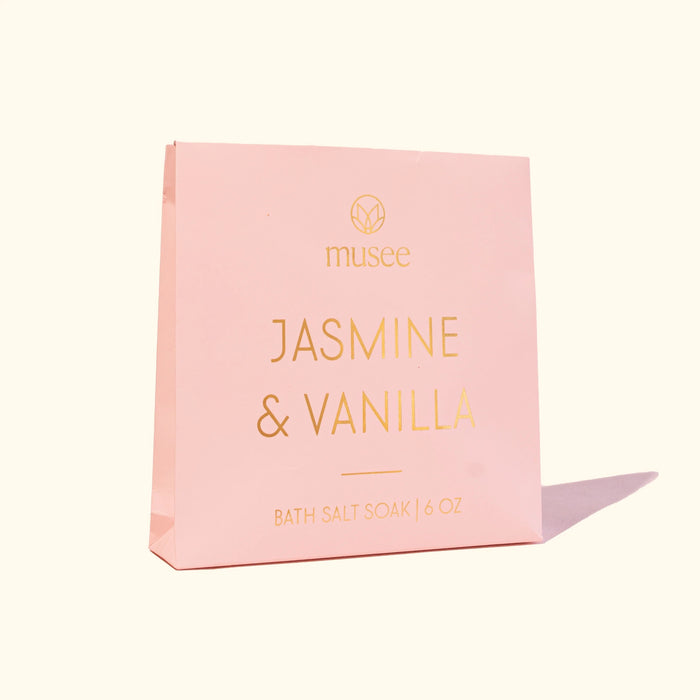 A pink package labeled “MUSEE - JASMINE & VANILLA MINI BATH SALT SOAK” by MUSEE, with a small pile of white bath salts spilled next to it, highlights the soothing benefits of natural bath products. Relax and unwind as you enjoy the calming jasmine vanilla aroma in your Epsom salt bath for ultimate tranquility.