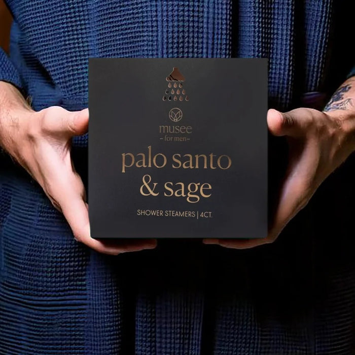 A box labeled “MUSEE - PALO SANTO & SAGE SHOWER STEAMERS” by MUSEE sits next to four white, round steamers featuring an elegant leaf design. The sleek black box with gold lettering is slightly open to reveal its illustrated inner packaging. This specialty MUSEE sage shower steamer elevates your daily routine, turning each shower into a luxurious aromatherapy experience.