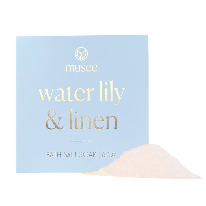 A blue box labeled “MUSEE - WATER LILY & LINEN MINI BATH SALT SOAK” by MUSEE sits neatly, with a small heap of bath salts—a calming blend of natural ingredients—spilled in front, inviting relaxation.