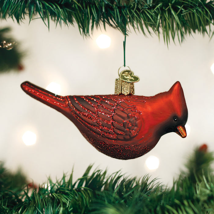 A red cardinal bird ornament with glittery details hangs from a Christmas tree branch, surrounded by green pine needles and soft, blurred holiday lights in the background.