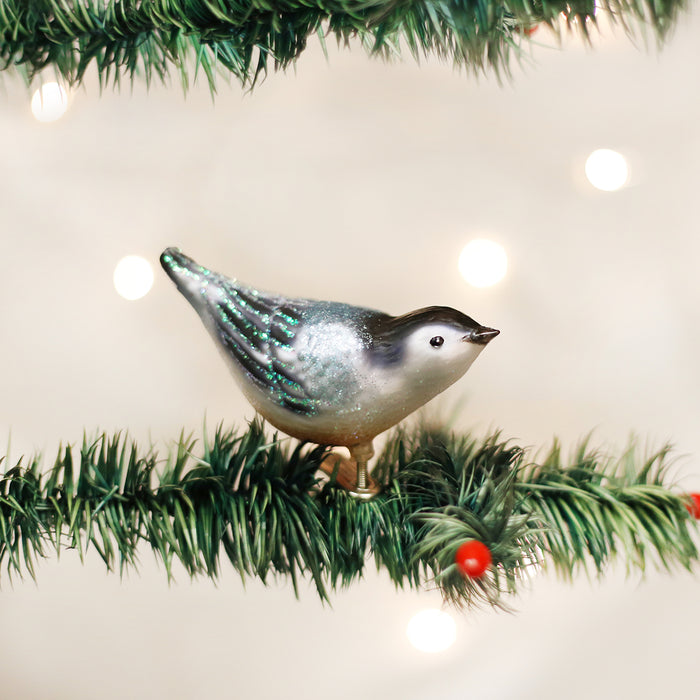 A shiny glass bird ornament with green and silver accents is clipped to an artificial pine branch, decorated with small red berries and surrounded by soft, glowing lights.