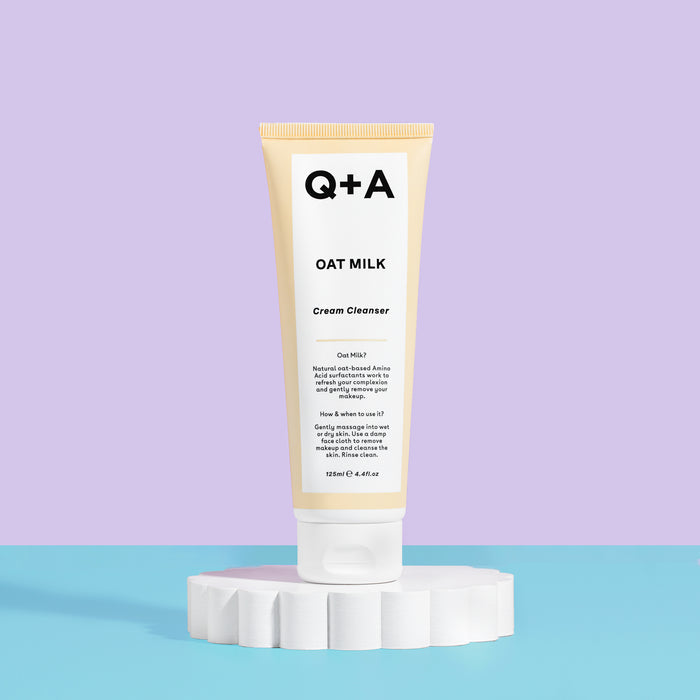 A tube of Q+A Oat Milk Cream Cleanser standing upright on a white pedestal against a split lavender and light blue background.