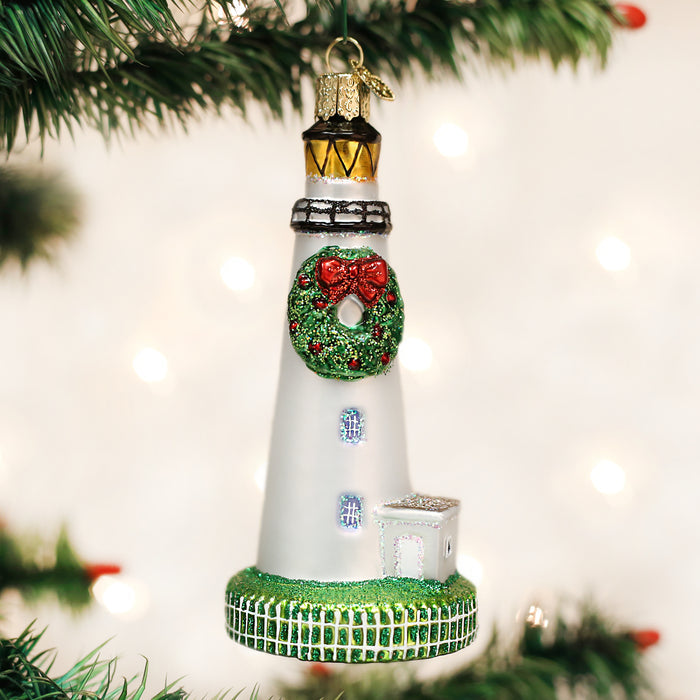 A Christmas ornament shaped like a white lighthouse with a wreath and red bow hangs from a tree branch, surrounded by green pine needles and blurred lights in the background.
