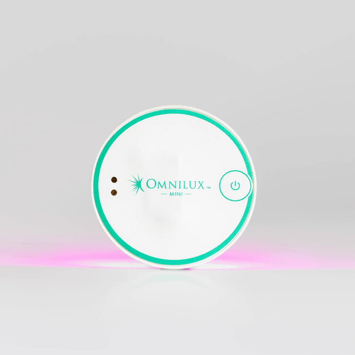 A round, white OMNILUX - BLEMISH ERASER KIT device from OMNILUX features a teal rim, power button, and logo; a soft pink light glows from underneath, showcasing its advanced blue and red light therapy technology.