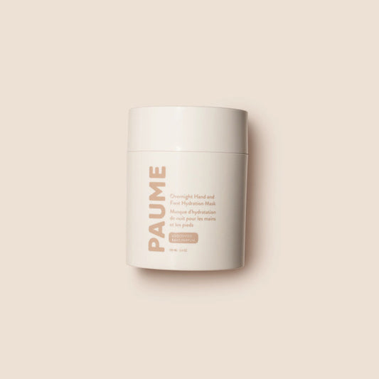 A white cylindrical container of PAUME’s OVERNIGHT HAND AND FOOT HYDRATION MASK rests on a creamy textured surface, with nourishing mask—ideal for dry, cracked skin—smeared around it.