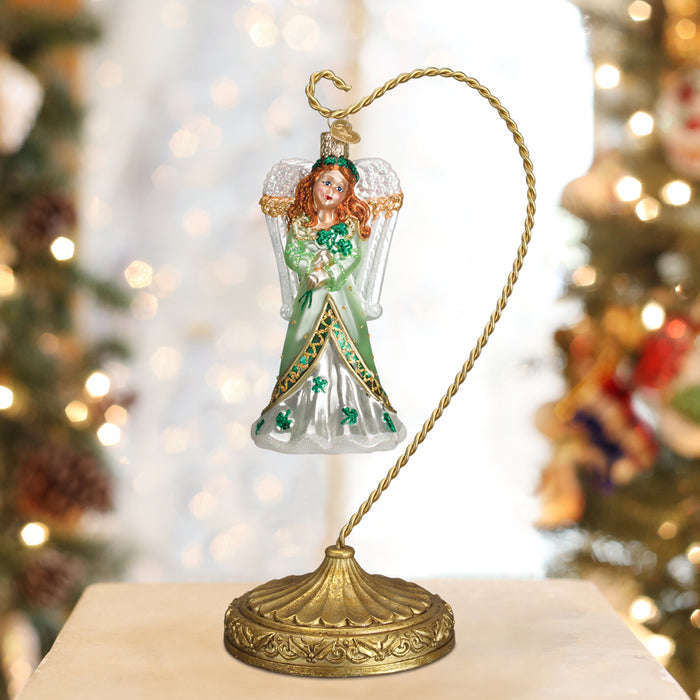 A glass angel ornament with red hair, a green and white dress decorated with shamrocks, and silver wings hangs from an ornate gold stand. Blurred Christmas tree lights are visible in the background.
