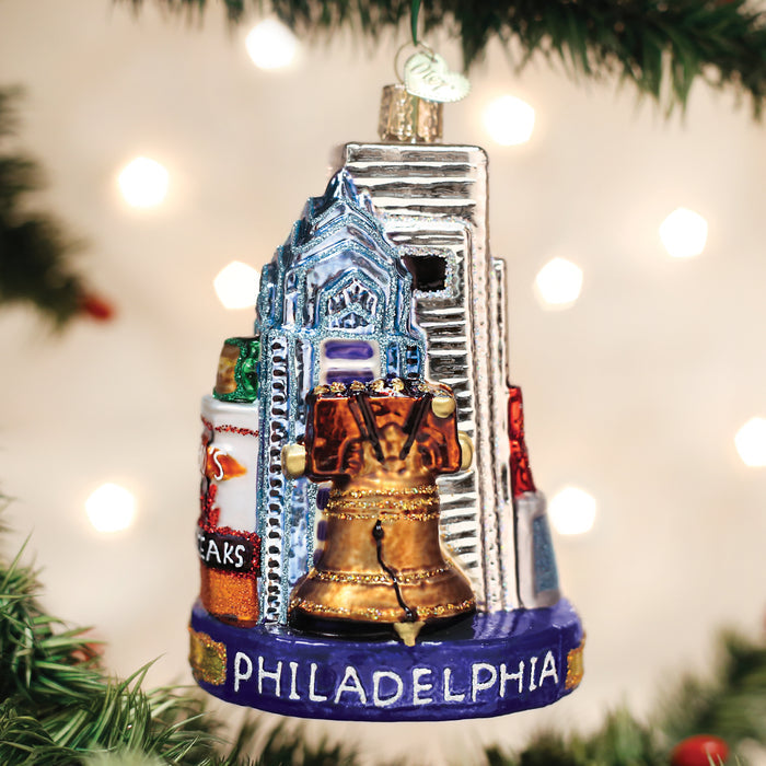 A Christmas tree ornament featuring Philadelphia landmarks, including the Liberty Bell, skyscrapers, and cheesesteaks, with PHILADELPHIA written at the base, hanging among green tree branches and blurred lights.