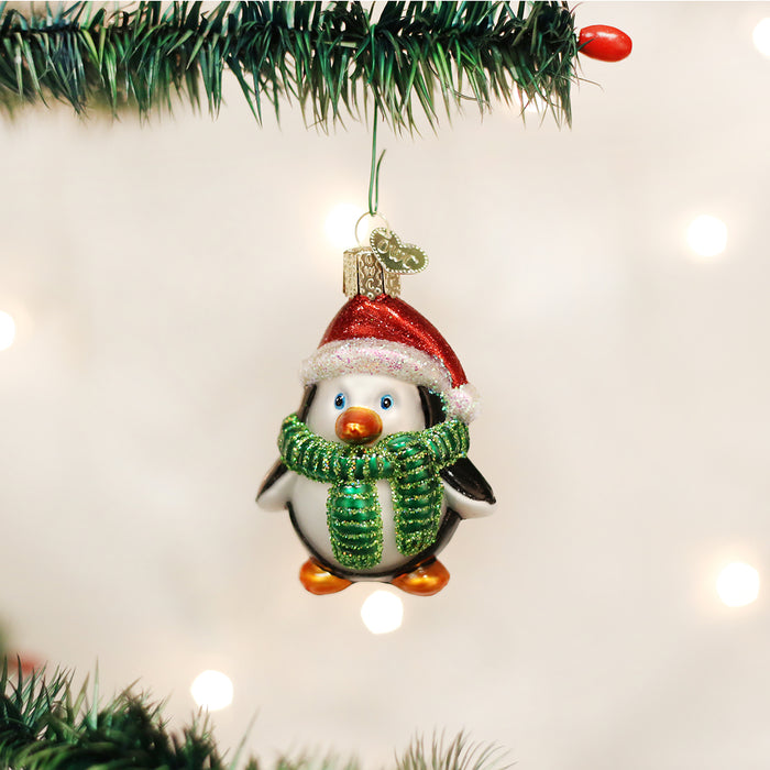 A Christmas tree ornament shaped like a penguin wearing a red Santa hat and a green scarf hangs from a branch, with festive lights glowing softly in the background.