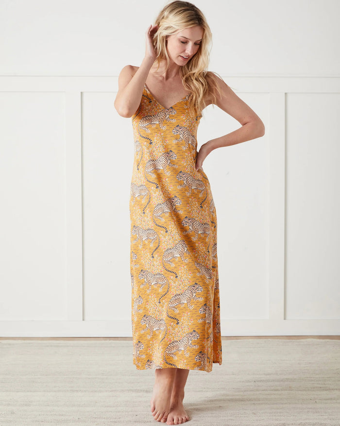 A woman stands barefoot indoors, wearing the PRINTFRESH Bagheera Eco Satin Chic to Sleep Nightie in Tobacco—a long, cheetah-patterned nightgown that captures the effortless elegance of sustainable sleepwear. She is looking down and touching her hair with one hand.