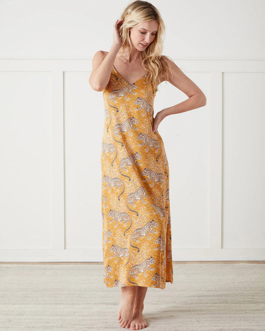 A woman stands barefoot indoors, wearing the PRINTFRESH Bagheera Eco Satin Chic to Sleep Nightie in Tobacco—a long, cheetah-patterned nightgown that captures the effortless elegance of sustainable sleepwear. She is looking down and touching her hair with one hand.