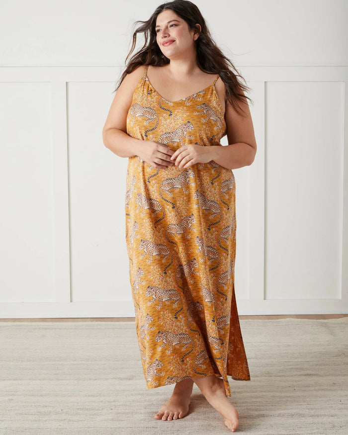 A woman stands barefoot indoors, wearing the PRINTFRESH Bagheera Eco Satin Chic to Sleep Nightie in Tobacco—a long, cheetah-patterned nightgown that captures the effortless elegance of sustainable sleepwear. She is looking down and touching her hair with one hand.