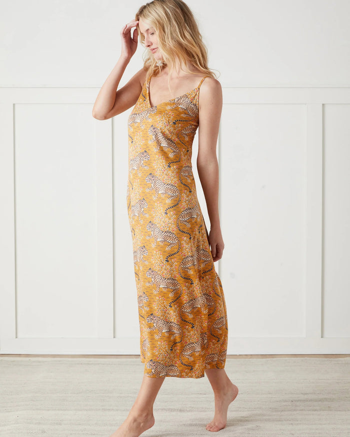 A woman stands barefoot indoors, wearing the PRINTFRESH Bagheera Eco Satin Chic to Sleep Nightie in Tobacco—a long, cheetah-patterned nightgown that captures the effortless elegance of sustainable sleepwear. She is looking down and touching her hair with one hand.