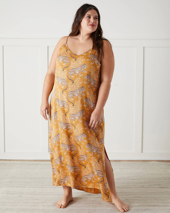 A woman stands barefoot indoors, wearing the PRINTFRESH Bagheera Eco Satin Chic to Sleep Nightie in Tobacco—a long, cheetah-patterned nightgown that captures the effortless elegance of sustainable sleepwear. She is looking down and touching her hair with one hand.