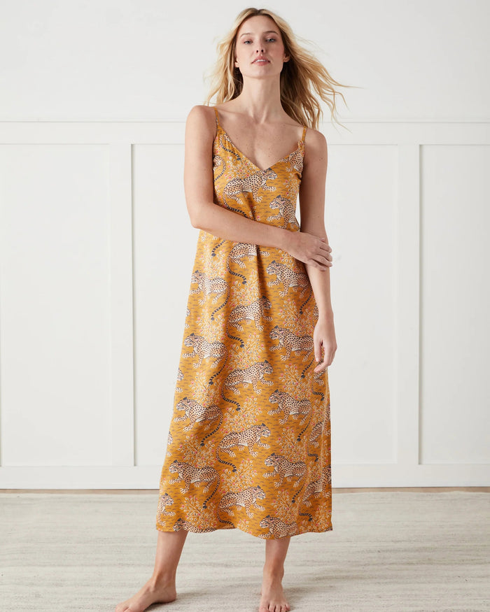 A woman stands barefoot indoors, wearing the PRINTFRESH Bagheera Eco Satin Chic to Sleep Nightie in Tobacco—a long, cheetah-patterned nightgown that captures the effortless elegance of sustainable sleepwear. She is looking down and touching her hair with one hand.