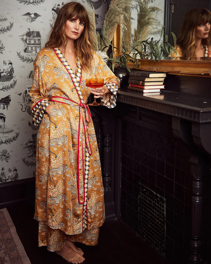 A woman stands barefoot, smiling, and wearing the PRINTFRESH Bagheera Eco Satin Robe in Tobacco—an eco-friendly viscose robe featuring a loose fit, bold patterns, and geometric trim. She is set against a white wooden panel wall with a beige carpet background.