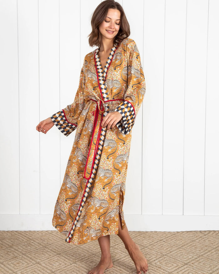 A woman stands barefoot by a dark fireplace, holding a drink, wearing the PRINTFRESH - BAGHEERA ECO SATIN ROBE IN TOBACCO from PRINTFRESH. Books and pampas grass adorn the mantel, while her reflection appears in the mirror behind her.