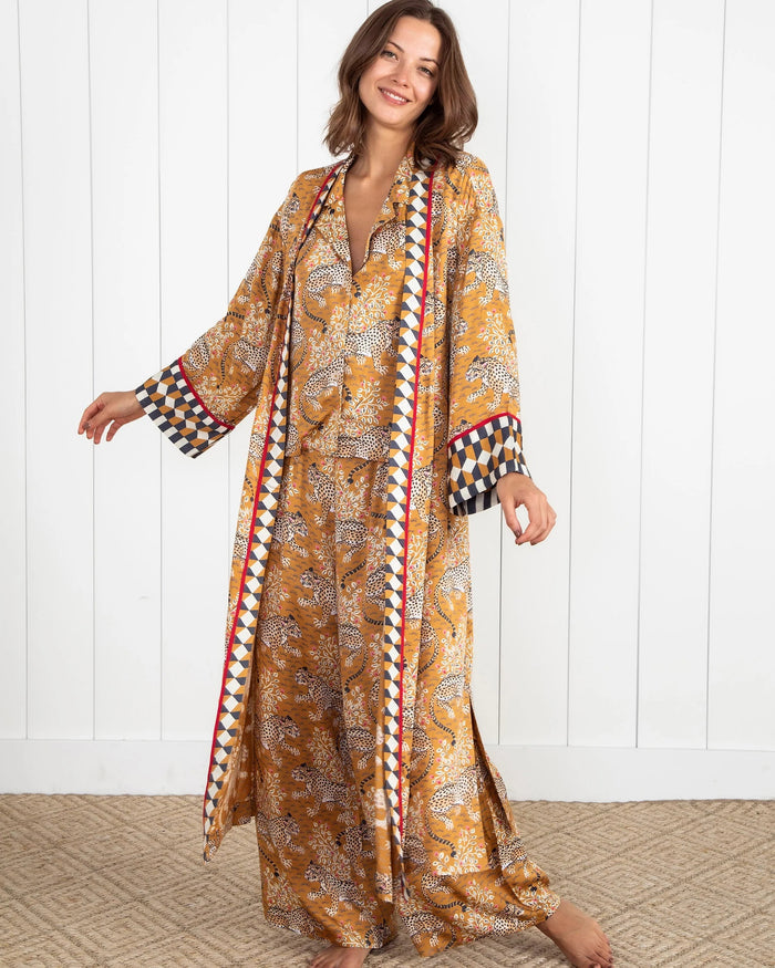 A woman stands barefoot by a dark fireplace, holding a drink, wearing the PRINTFRESH - BAGHEERA ECO SATIN ROBE IN TOBACCO from PRINTFRESH. Books and pampas grass adorn the mantel, while her reflection appears in the mirror behind her.