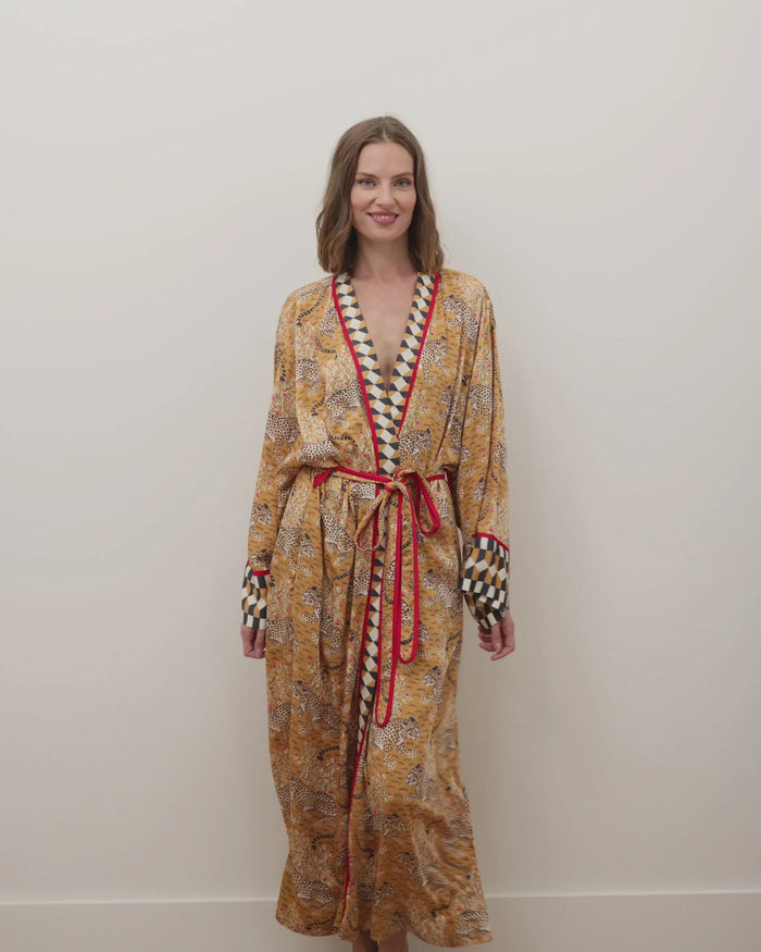 A woman stands barefoot by a dark fireplace, holding a drink, wearing the PRINTFRESH - BAGHEERA ECO SATIN ROBE IN TOBACCO from PRINTFRESH. Books and pampas grass adorn the mantel, while her reflection appears in the mirror behind her.