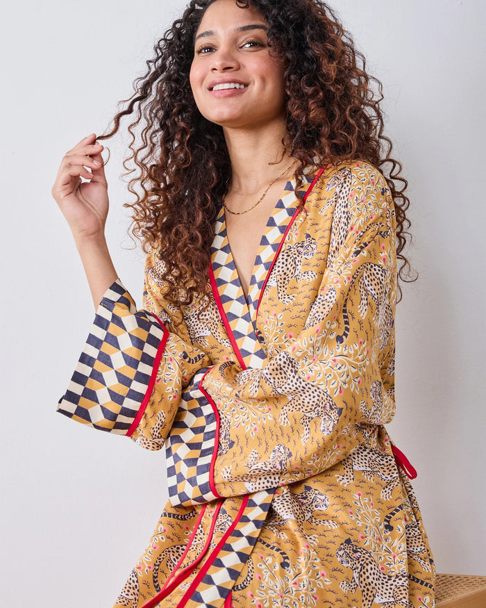 A woman stands barefoot by a dark fireplace, holding a drink, wearing the PRINTFRESH - BAGHEERA ECO SATIN ROBE IN TOBACCO from PRINTFRESH. Books and pampas grass adorn the mantel, while her reflection appears in the mirror behind her.