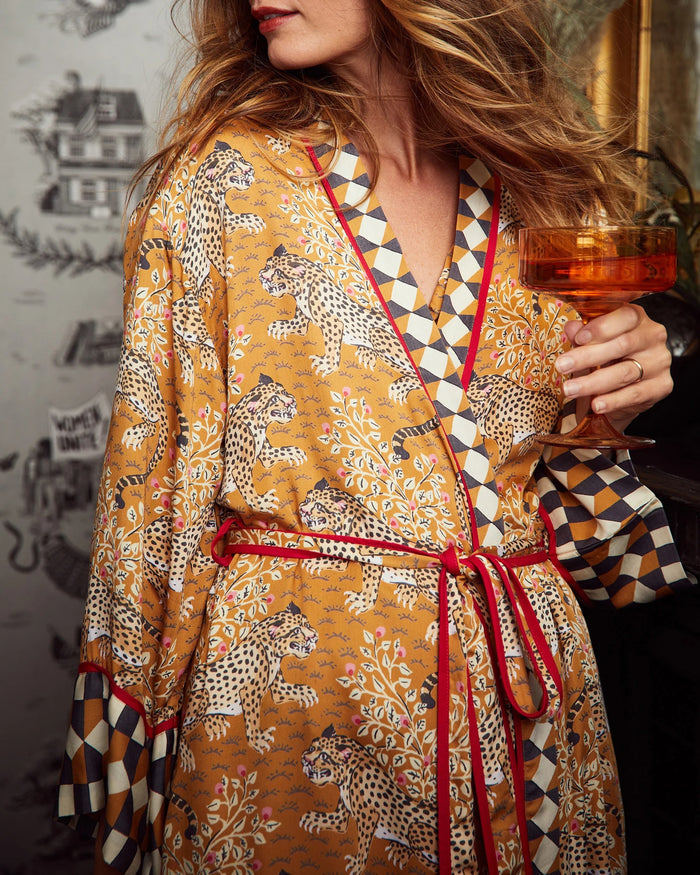 A woman stands barefoot by a dark fireplace, holding a drink, wearing the PRINTFRESH - BAGHEERA ECO SATIN ROBE IN TOBACCO from PRINTFRESH. Books and pampas grass adorn the mantel, while her reflection appears in the mirror behind her.