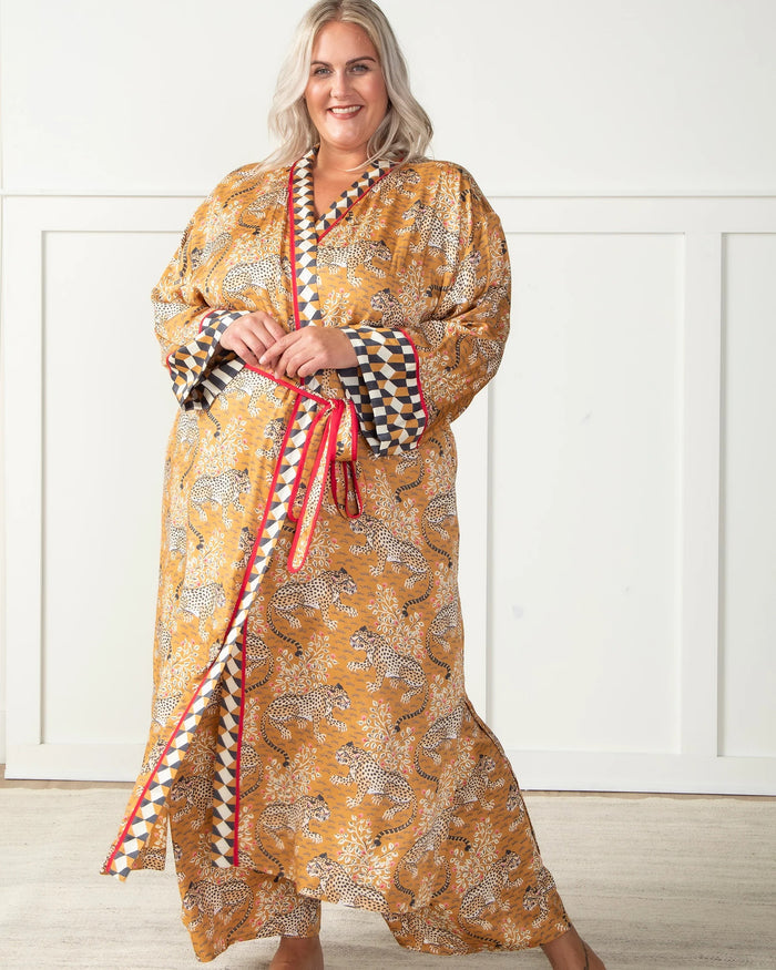 A woman stands barefoot by a dark fireplace, holding a drink, wearing the PRINTFRESH - BAGHEERA ECO SATIN ROBE IN TOBACCO from PRINTFRESH. Books and pampas grass adorn the mantel, while her reflection appears in the mirror behind her.