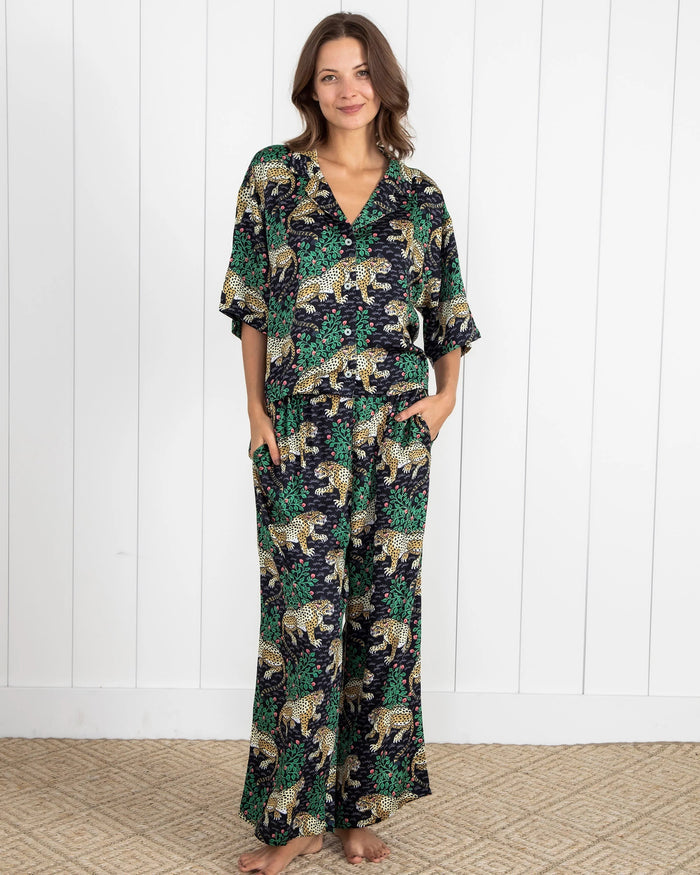 A woman wearing the PRINTFRESH - BAGHEERA SATIN WILDEST DREAM PAJAMA SET IN INK sits relaxed in a yellow armchair on a wooden floor, with one arm resting on the chair and one leg extended, sporting matching PRINTFRESH slippers with black fur trim.