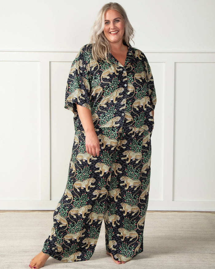 A woman stands barefoot on a woven rug in front of a white paneled wall, wearing the PRINTFRESH - Bagheera Satin Wildest Dream Pajama Set in Ink from PRINTFRESH, featuring a vibrant leopard and foliage pattern with wide-leg pants.