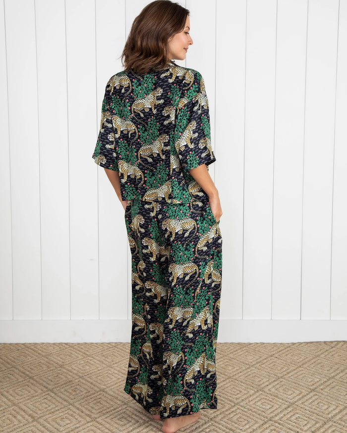 A woman stands barefoot on a woven rug in front of a white paneled wall, wearing the PRINTFRESH - Bagheera Satin Wildest Dream Pajama Set in Ink from PRINTFRESH, featuring a vibrant leopard and foliage pattern with wide-leg pants.