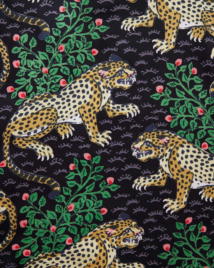 A woman stands barefoot on a woven rug in front of a white paneled wall, wearing the PRINTFRESH - Bagheera Satin Wildest Dream Pajama Set in Ink from PRINTFRESH, featuring a vibrant leopard and foliage pattern with wide-leg pants.