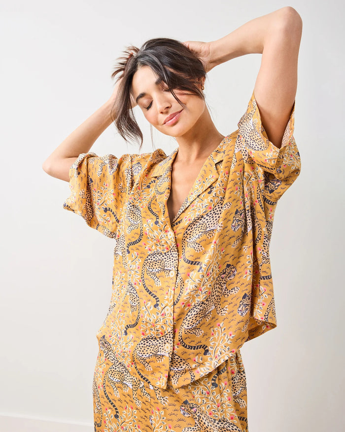 A woman in the PRINTFRESH - Bagheera Satin Wildest Dream Pajama Set in Tobacco sits barefoot on a blue velvet fringe-trimmed couch, flanked by teal geometric pillows. Blue curtains and patterned wallpaper provide a backdrop, creating a cozy ambiance that reflects the eco-friendly elegance of this PRINTFRESH pajama set.