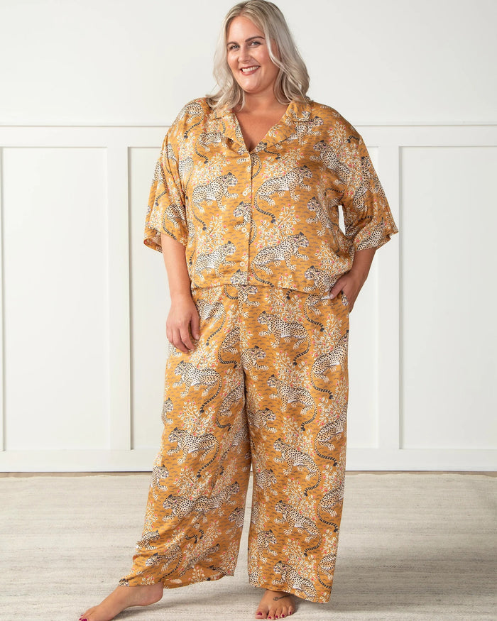 A woman in the PRINTFRESH - Bagheera Satin Wildest Dream Pajama Set in Tobacco sits barefoot on a blue velvet fringe-trimmed couch, flanked by teal geometric pillows. Blue curtains and patterned wallpaper provide a backdrop, creating a cozy ambiance that reflects the eco-friendly elegance of this PRINTFRESH pajama set.