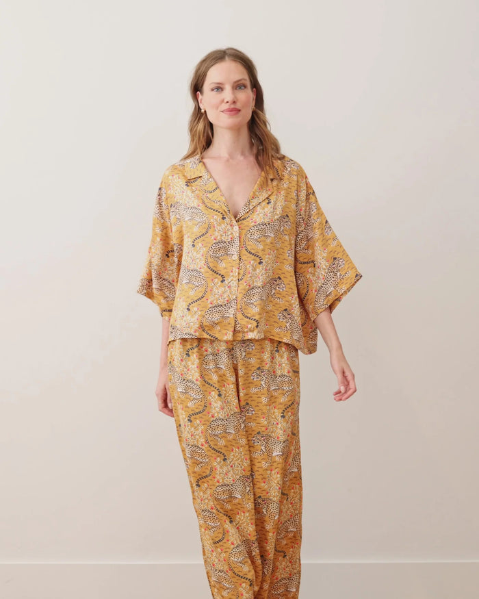 Wearing the PRINTFRESH - Bagheera Satin Wildest Dream Pajama Set in Tobacco, a woman stands indoors with a smile, posing with one arm raised. A lamp, books, and abstract art decorate the background.